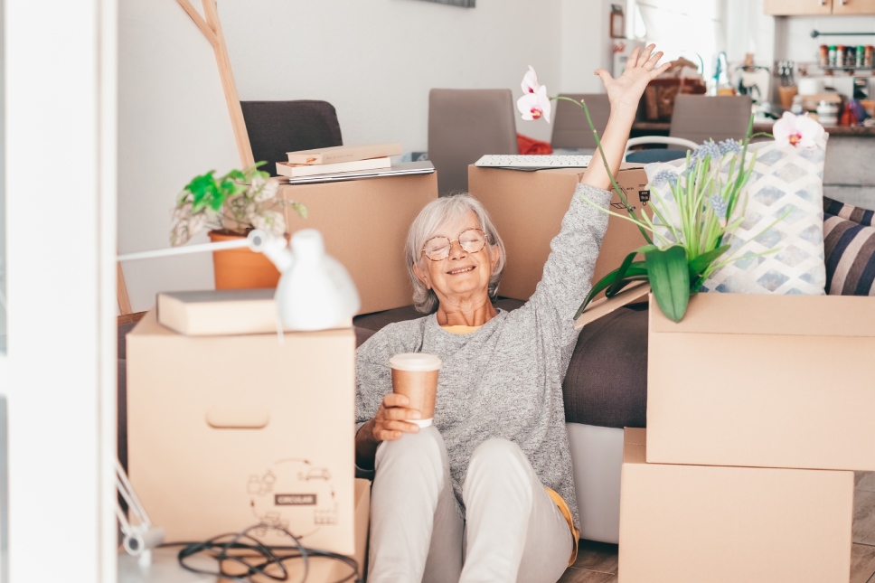 Happy senior woman involved in moving house sitting among cardboard boxes takes a break with a coffee, concept of moving, retirement, new life, buying, renting, apartment, house