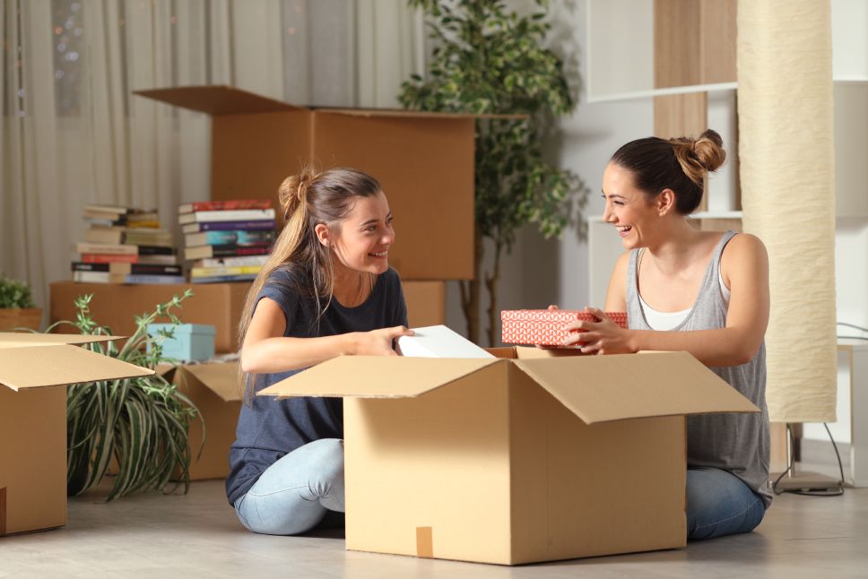 Two happy roommates talking unboxing gelongings moving home sitting on the floor