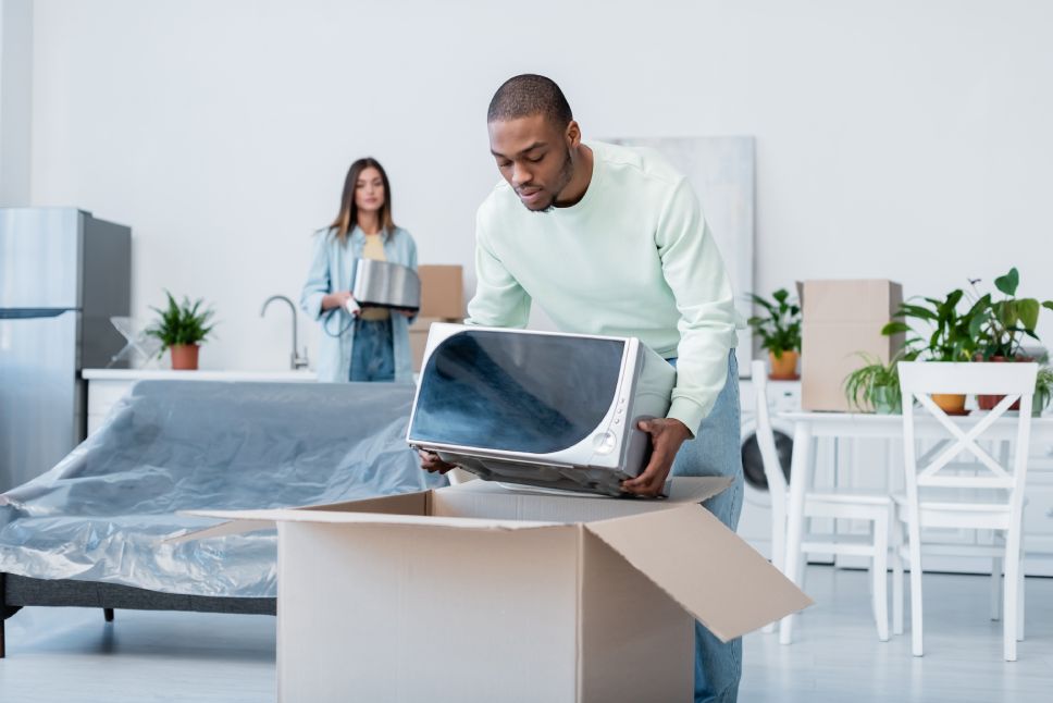 african american man putting microwave oven in carton box while packing during relocation