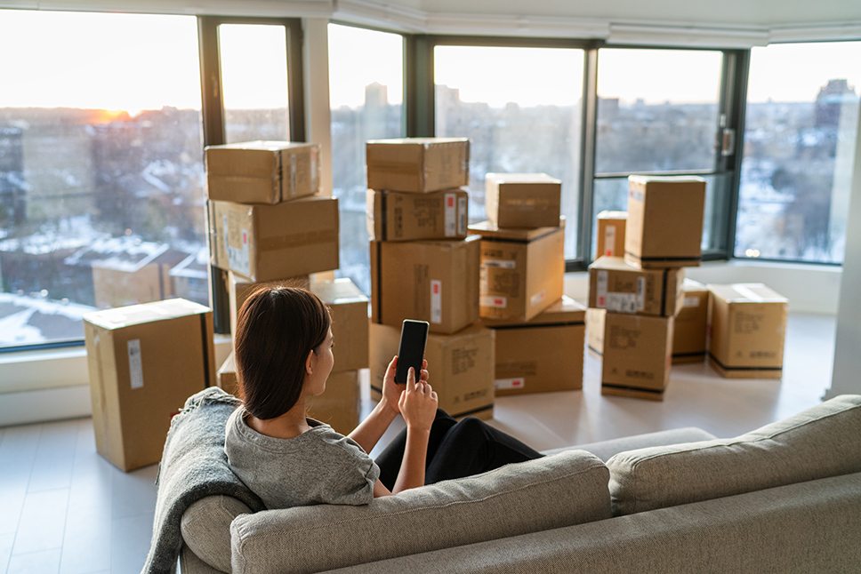 Woman on couch checking cell phone amidst moving boxes in apartment