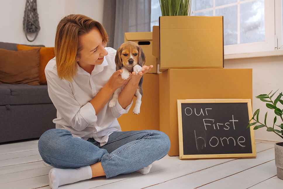 A woman sitting on the floor holding a small dog, surrounded by moving boxes