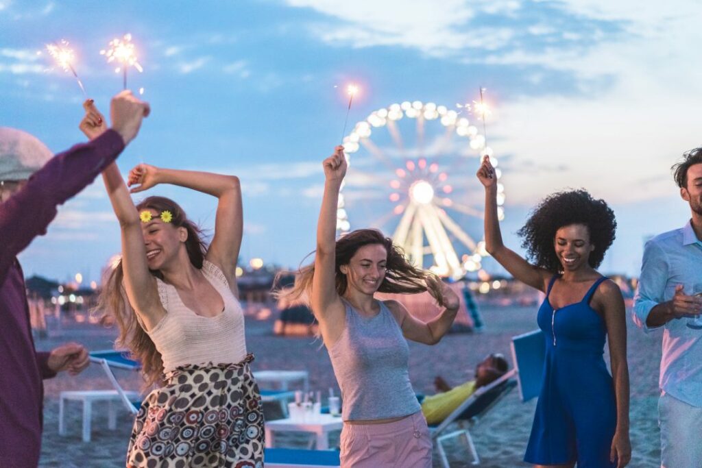 Happy friends making beach party after sunset with fireworks and drinking champagne - Young people having fun dancing outdoor  - Soft focus on center girl face - Youth and festival concept
