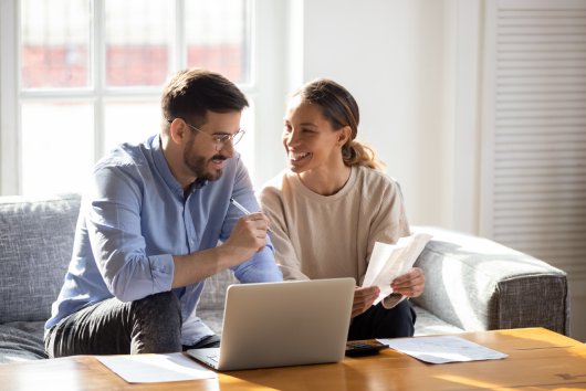 Happy young married couple sit on couch calculate expenses use easy online banking service at home