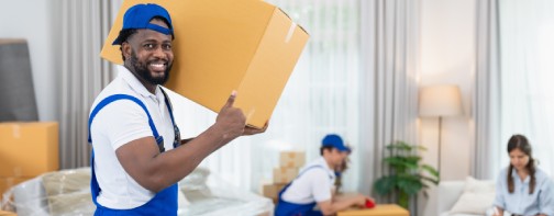 Smiling man mover worker in blue uniform carrying cardboard boxes moving to living room in new house. Professional delivery and moving service.