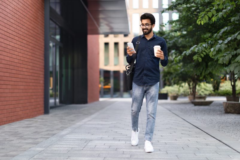 Cheerful millennial arab guy employee going to office in the morning, using phone on the street, drinking takeaway coffee. Happy middle eastern businessman checking email on smartphone, copy space