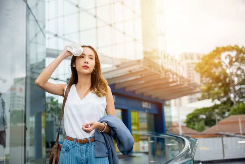 Angry female having sunstroke skin damage from sun UV city air pollution outside on street, Overheating Asian beautiful business woman drying sweat her face with cloth in warm summer day hot weather