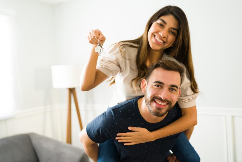 Handsome man piggybacking his girlfriend showing their new home keys after buying a house