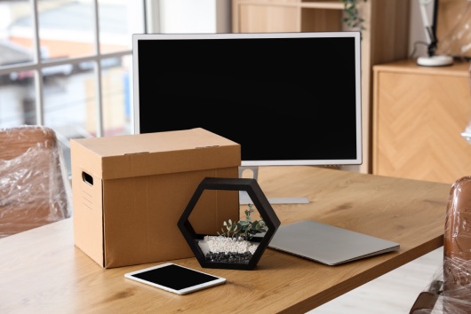 Desk with tablet computer, laptop, PC monitor and cardboard box in office on moving day