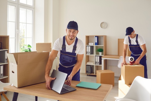 Young mover man packing laptop for moving, individual or company worker job moving office things, possessions, boxes, busy workmen professional team wearing uniform helping to change residence
