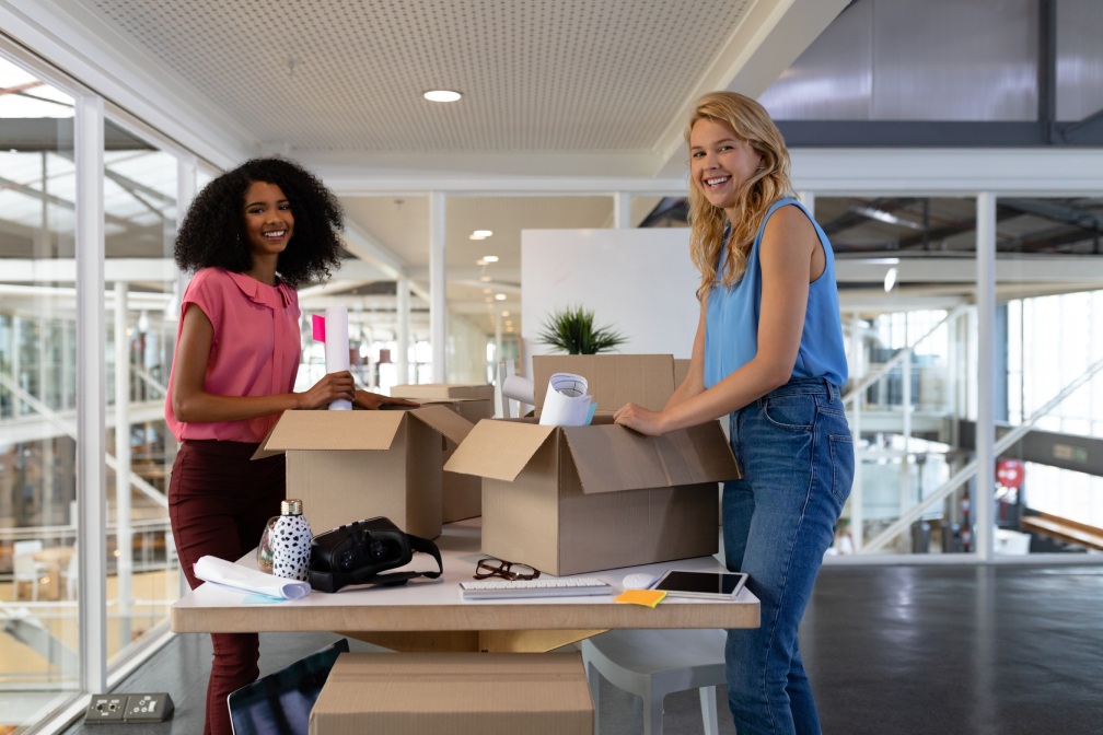 Portrait of young diverse female executives unpacking cardboard boxes in office. This is a casual creative start-up business office for a diverse team