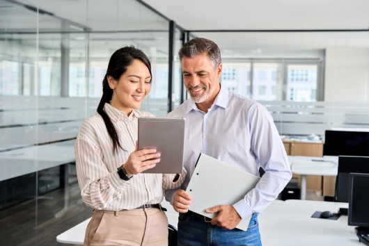Two happy professional business people team Asian woman and Latin man workers working using digital tablet tech discussing financial market data standing at corporate office meeting.