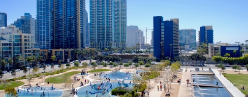 Panoramic view on a sunny day of children playing, people relaxing and cooling off in fountains in public park opposite port of San Diego, Southern California.