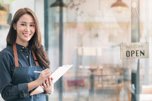 Beautiful asian young barista woman in apron holding clipboard and standing in front of the door of cafe with open sign board. Business owner startup concept.