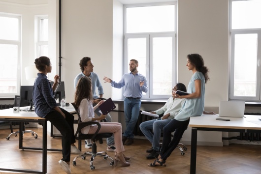 Multiethnic staff members gathered in coworking solve business, listen to male team leader during morning briefing in modern company office. Boss share information, corporate goals, tell about