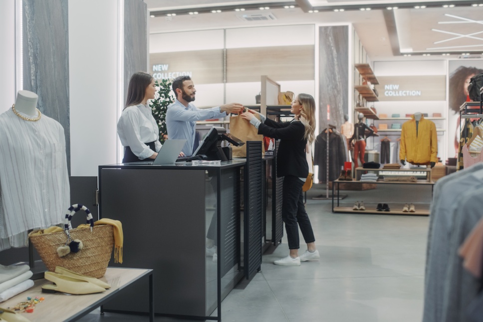 Clothing Store Checkout Cashier Counter: Beautiful Young Woman Buys Blouse from Friendly Retail Sales assistant, Receiving Bag with Purchases. Trendy Fashion Shop with of Designer Brands