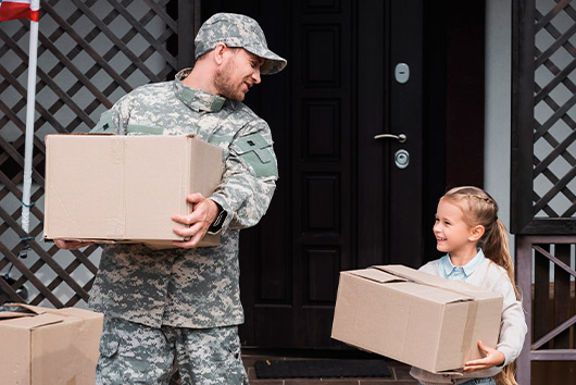 A soldier hands a box to a smiling little girl, symbolizing support and connection in a military community.