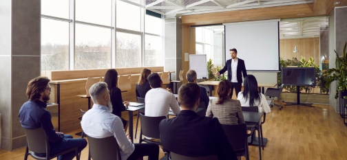 Team of people having class with business trainer. Group of male and female employees sitting at desks in modern office and listening to lecture by experienced teacher sharing knowledge and guidance