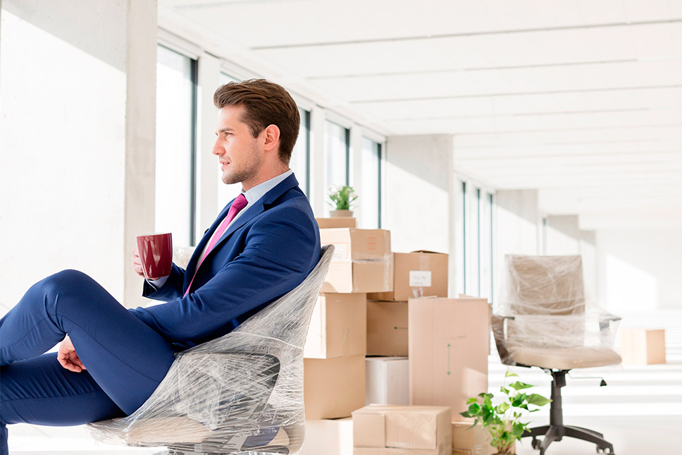 A suited man sits on a chair with boxes around him, representing the process of moving a corporate office in the Inland Empire.
