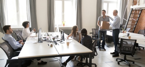 Office workers sitting on their places looking at boss ceo introduce a new employee to colleagues. Millennial man holds box has first workday in company getting vacancy feels satisfied and happy