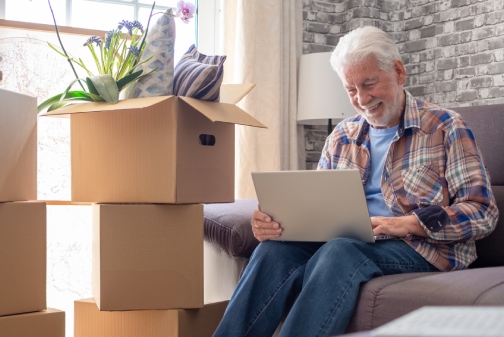 Happy senior man involved in moving house sitting among cardboard boxes using laptop, concept of relocating, retirement, new life, buying, renting, apartment, house