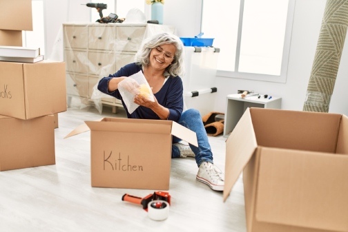 Middle age grey-haired woman smiling happy unboxing kitchen cardboard box at new home.