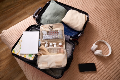 A suitcase is open on a bed, containing neatly arranged clothing, toiletries, and travel necessities. A notepad, headphones, and a smartphone are nearby, indicating preparation for a journey.