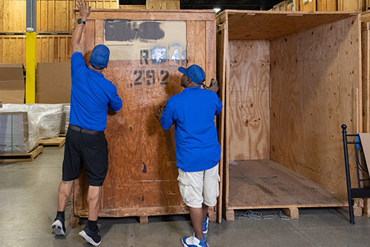 Two men wearing blue shirts are carrying a wooden box, illustrating long-term storage options in San Marcos. Oakley movers