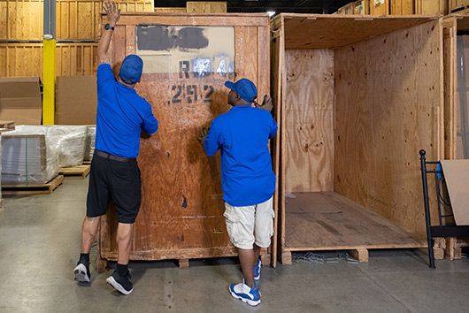 Two men wearing blue shirts are transporting a large wooden box, featured in the Winter Storage Guide for San Marcos Facilities.